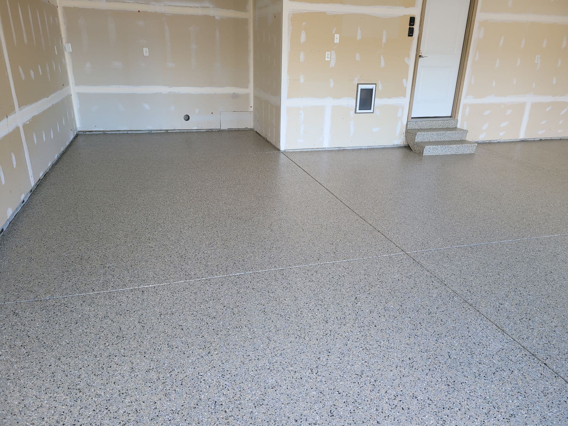 Empty garage interior with freshly coated speckled epoxy floor, smooth beige drywall, and white door with small concrete steps, conveying a clean, new feel.