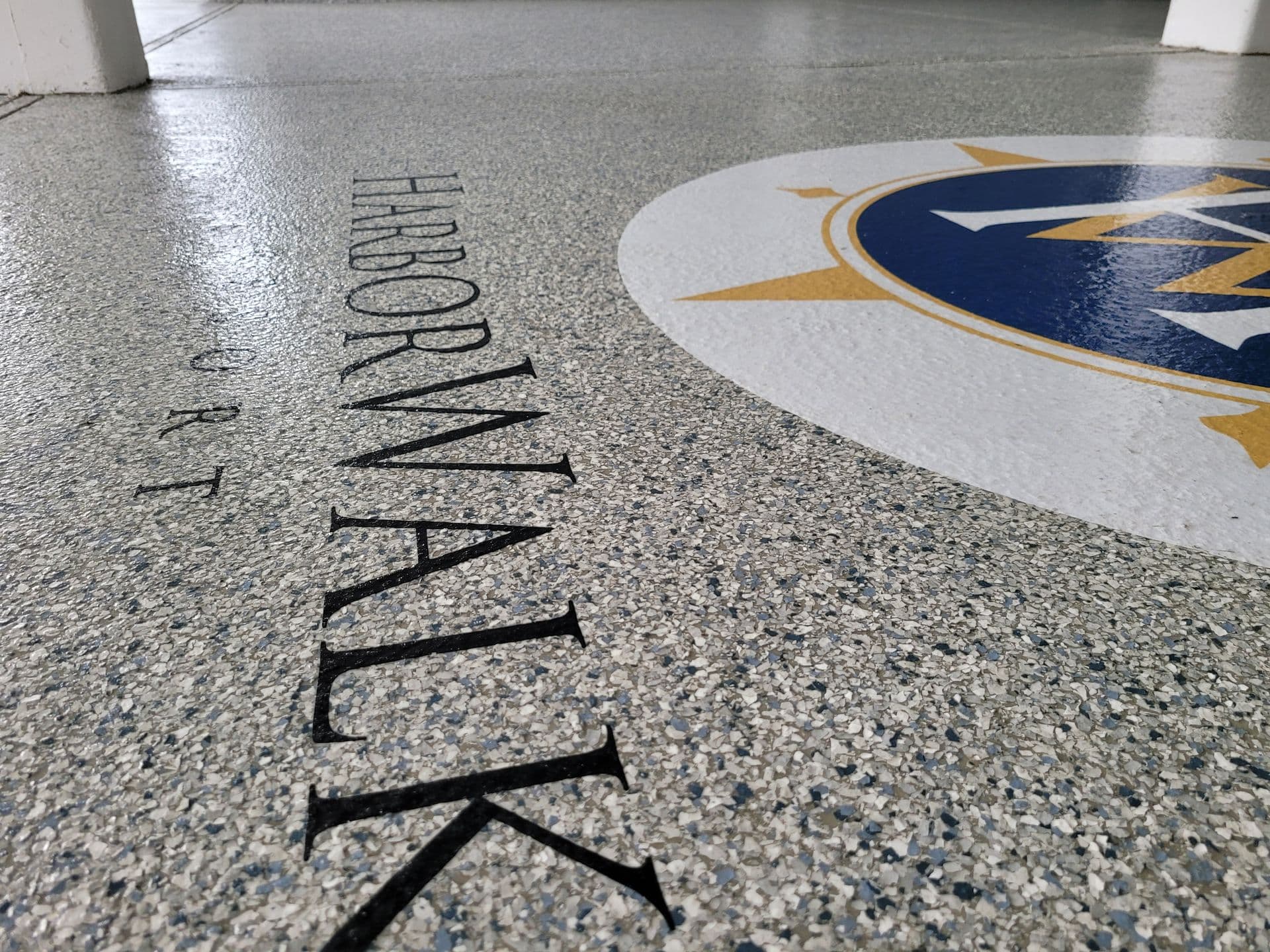 Close-up of a textured ground with the word "HARBORWALK" in black, beside a white circle featuring a blue and gold compass motif.