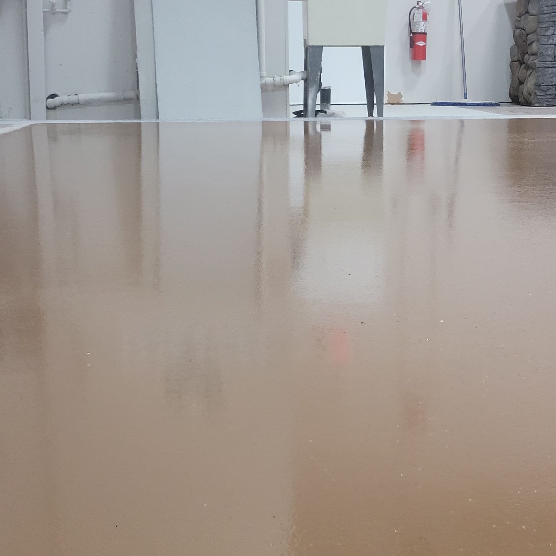 Smooth, shiny brown floor in an industrial setting, reflecting pipes and a fire extinguisher on a wall. Clean, polished appearance, minimalistic.