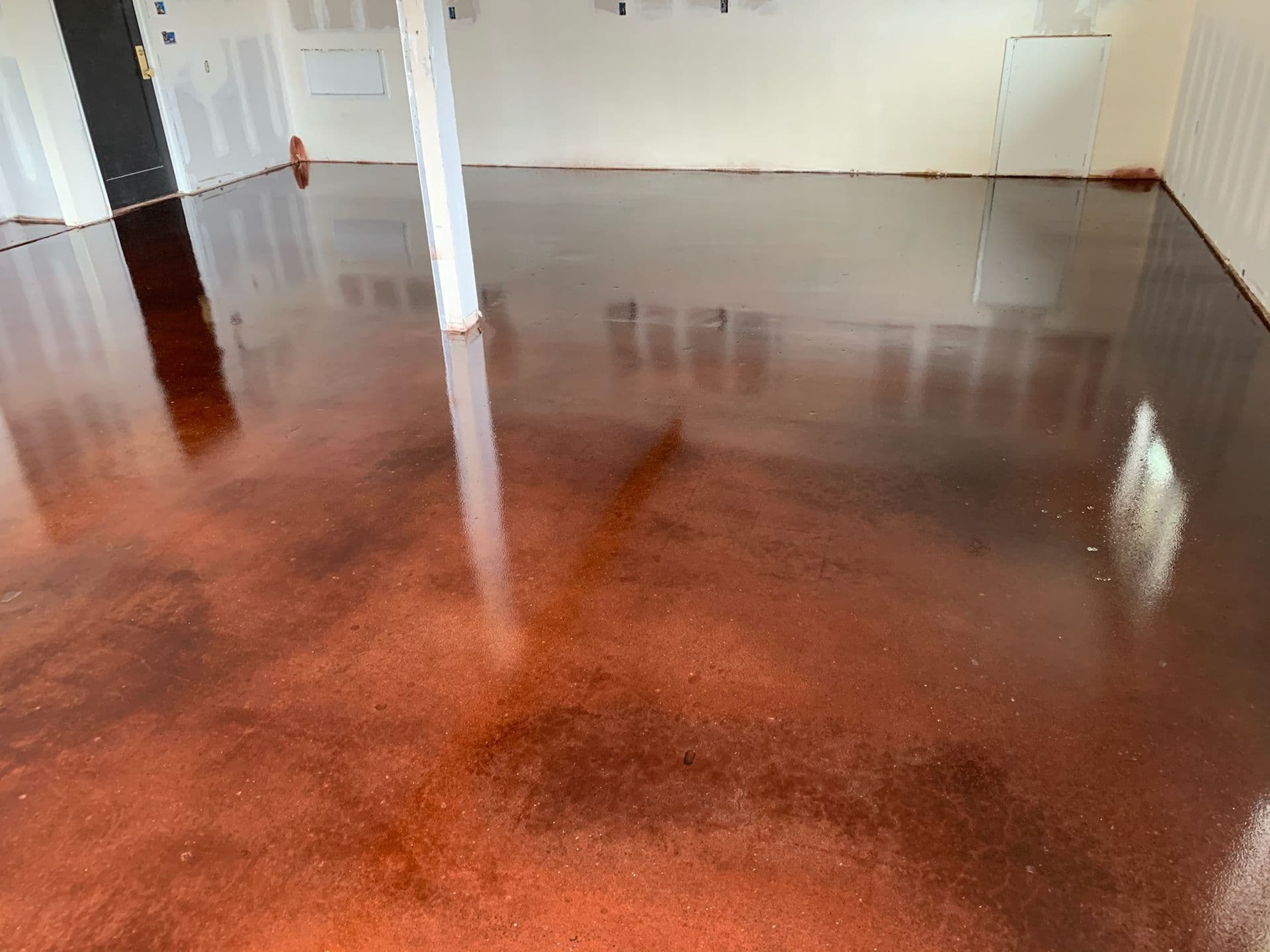 Smooth, polished brown concrete floor in an empty room with white walls and a white support column. The floor reflects light, creating a glossy effect.