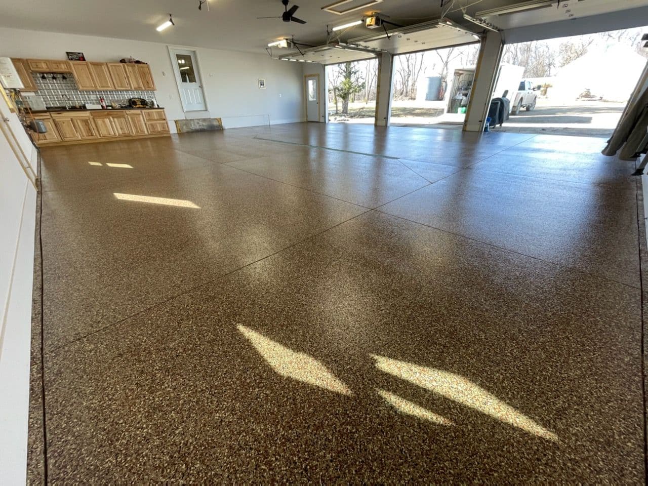 Spacious, sunlit garage with speckled epoxy floors, wooden cabinets, and open garage doors revealing an outdoor view. Clean and organized ambiance.