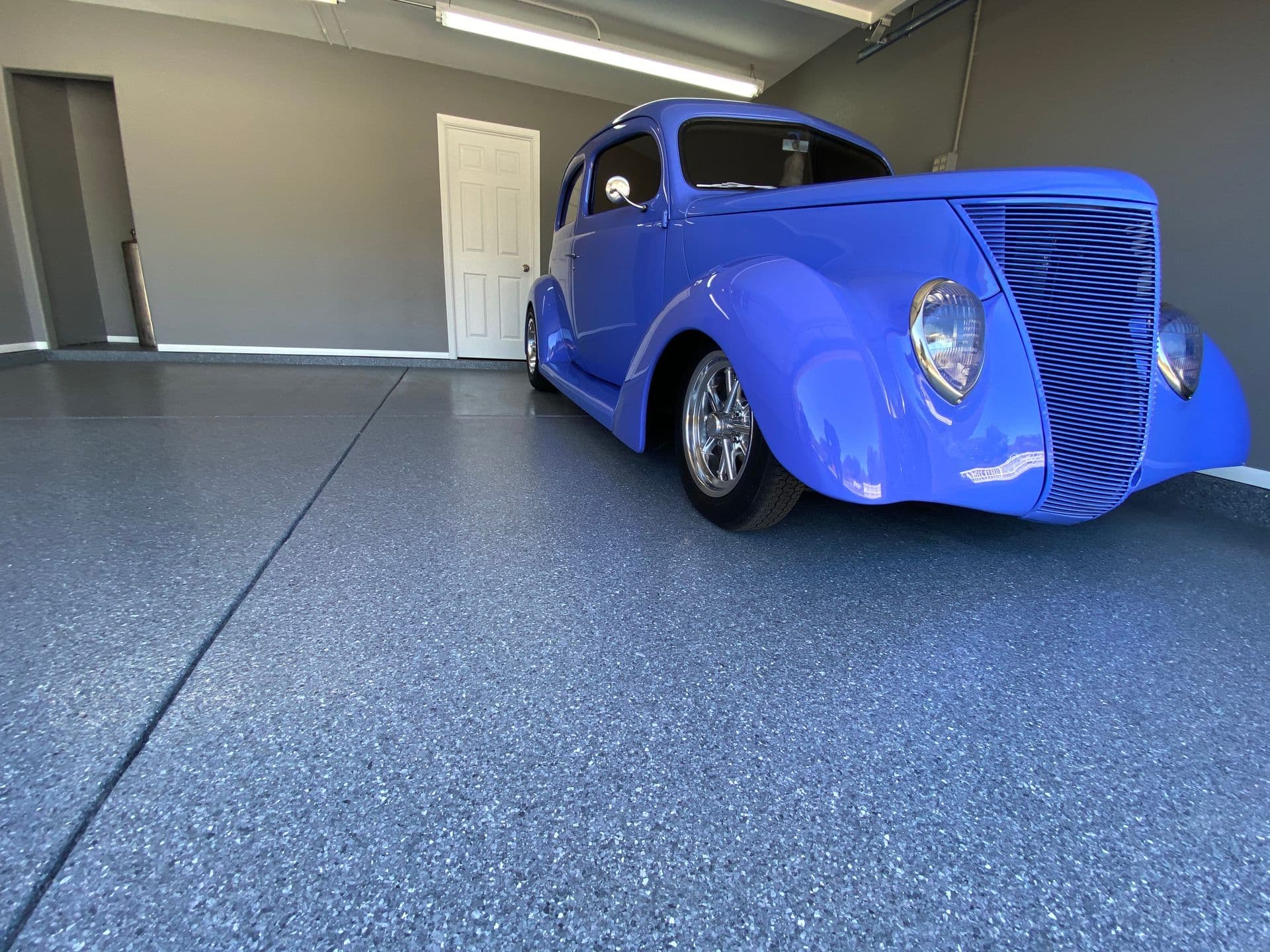 A vibrant blue vintage car with a sleek grille and chrome wheels is parked in a clean, gray-walled garage. The scene feels still and pristine.