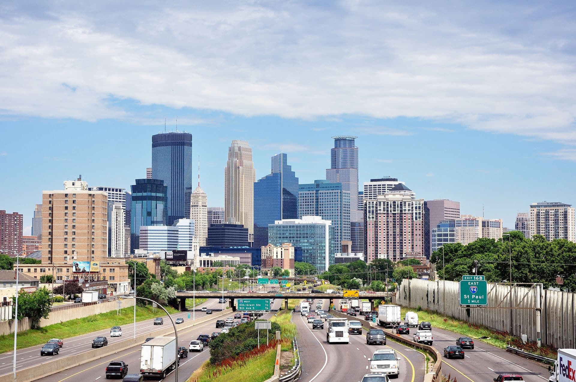 A bustling highway leads to a city skyline with modern skyscrapers under a bright blue sky. Greenery lines the road, adding a touch of nature to the urban scene.