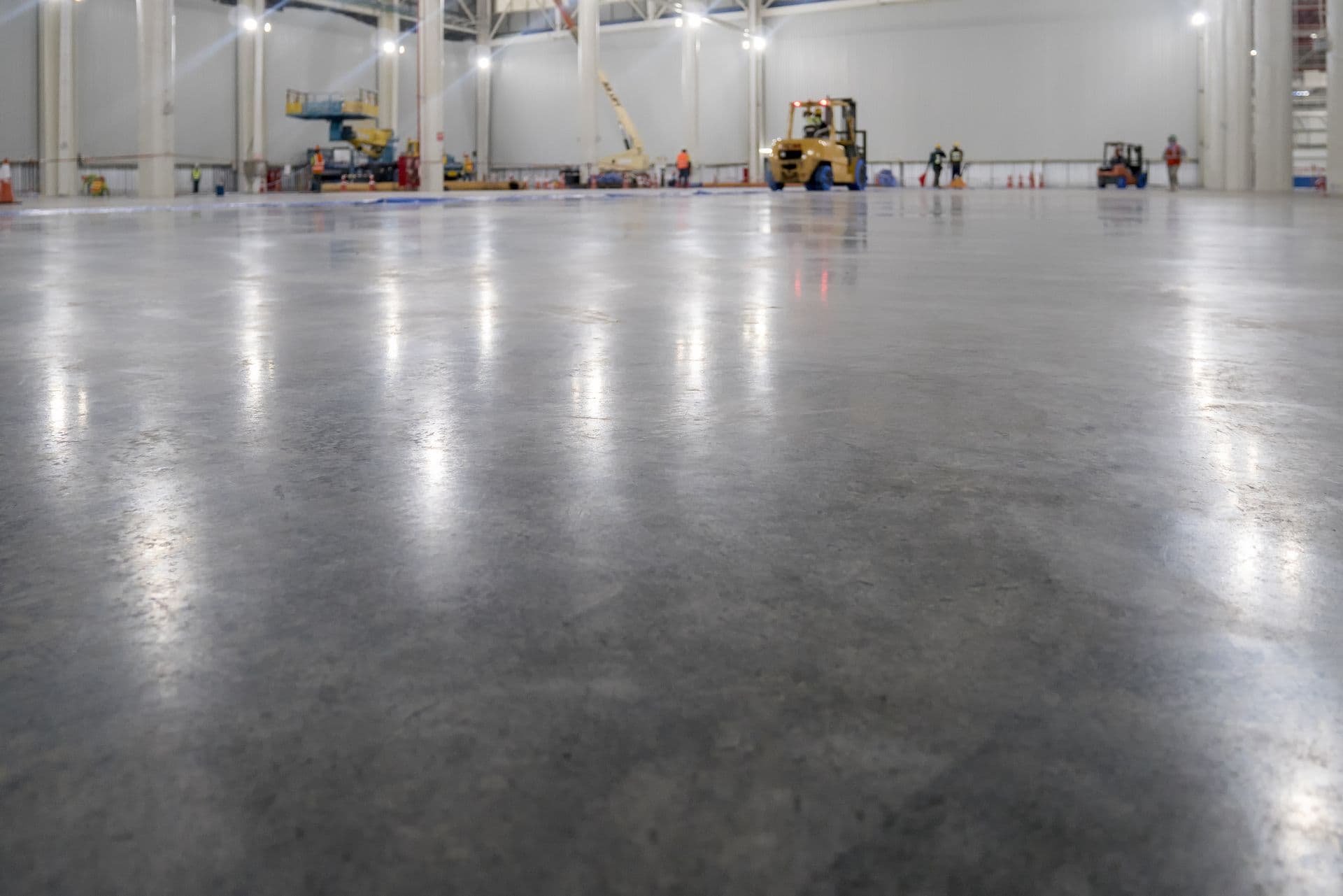 Wide shot of an empty, polished concrete floor in a large industrial warehouse. Workers and machinery are visible in the background. Bright, clean, and spacious.