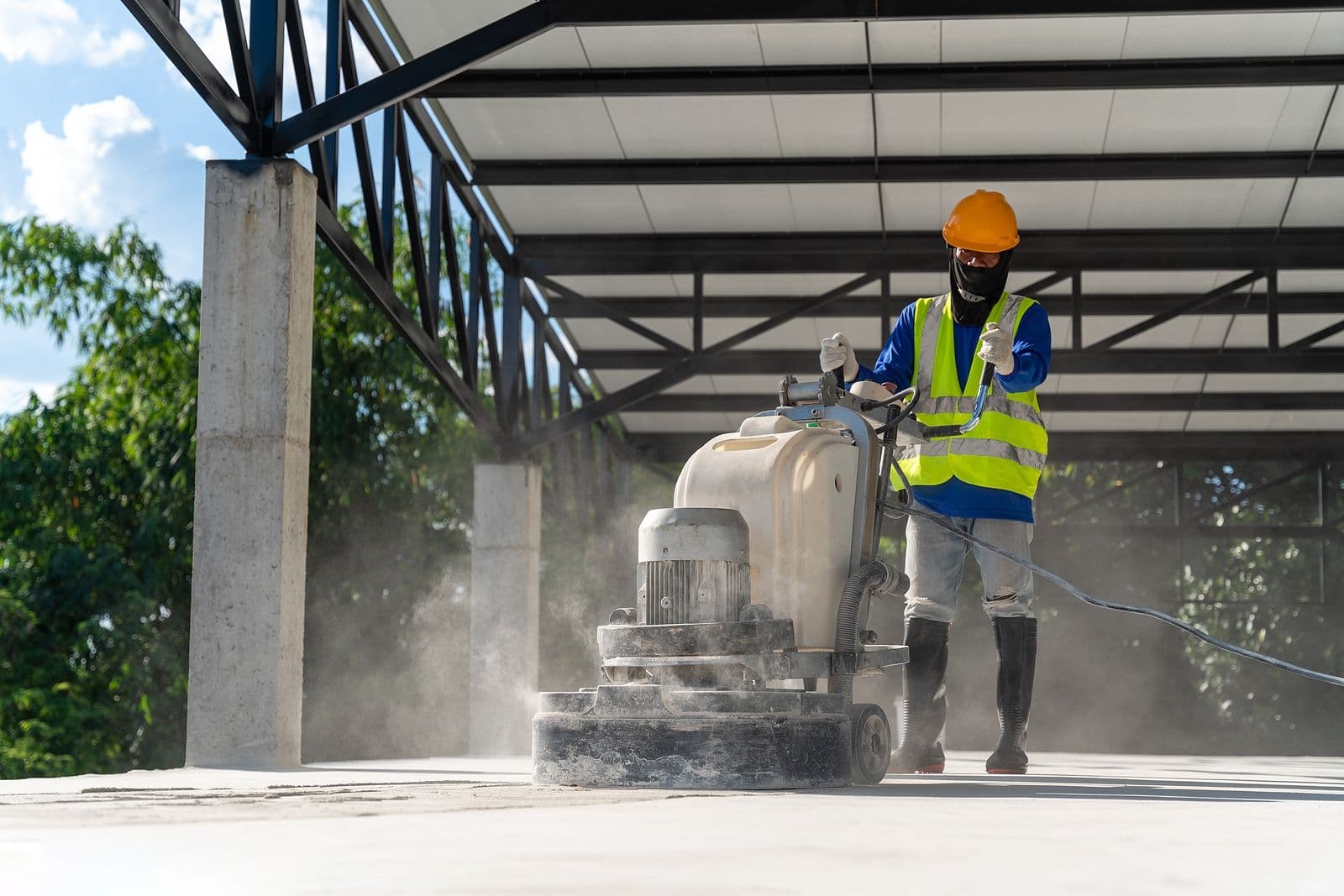 Construction worker operating concrete grinding machine under industrial structure, creating dust while performing surface preparation work