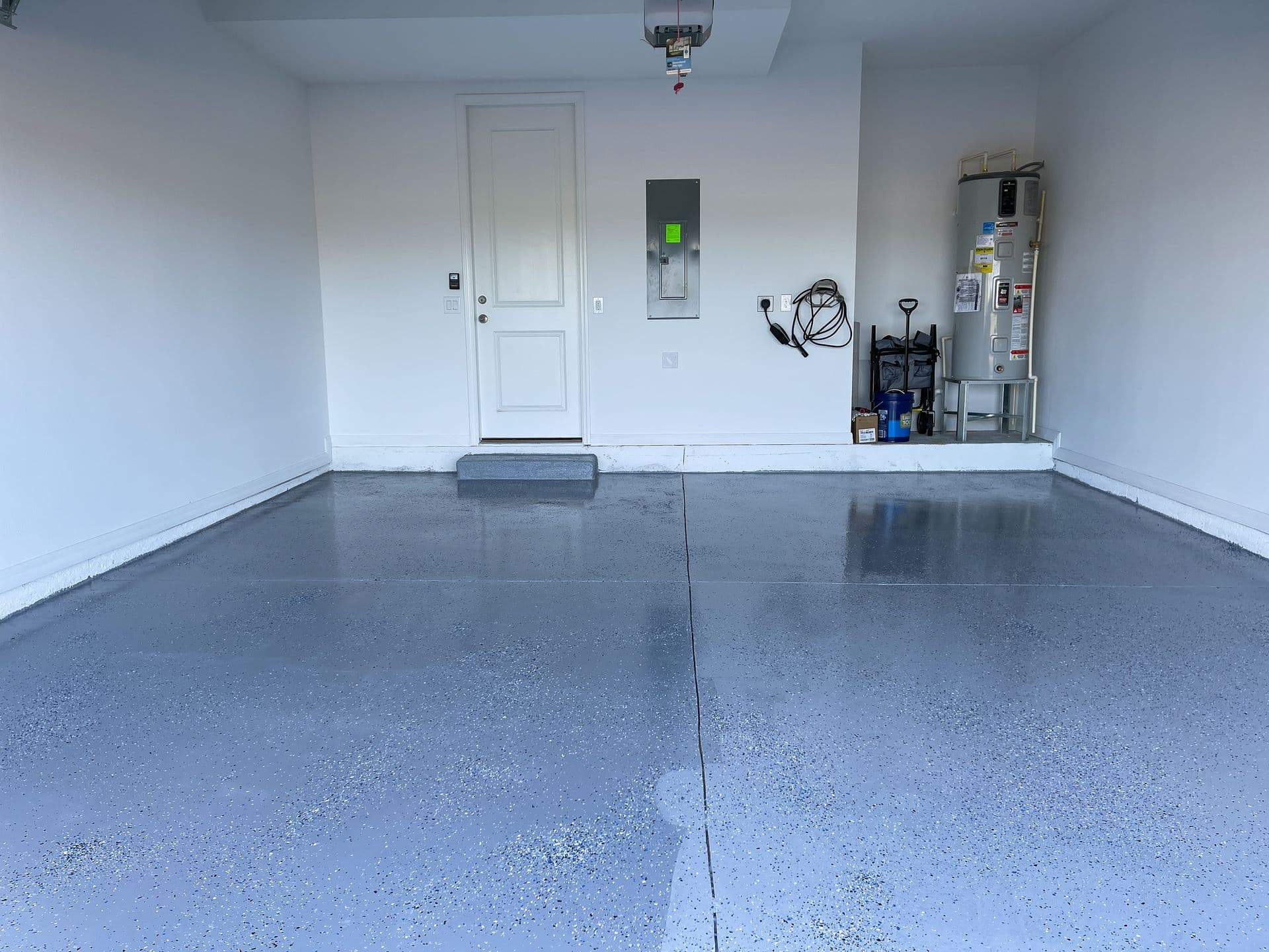 Empty garage with a smooth, speckled gray floor. White walls, a closed door, utility panel, water heater, and neatly stored cleaning items.