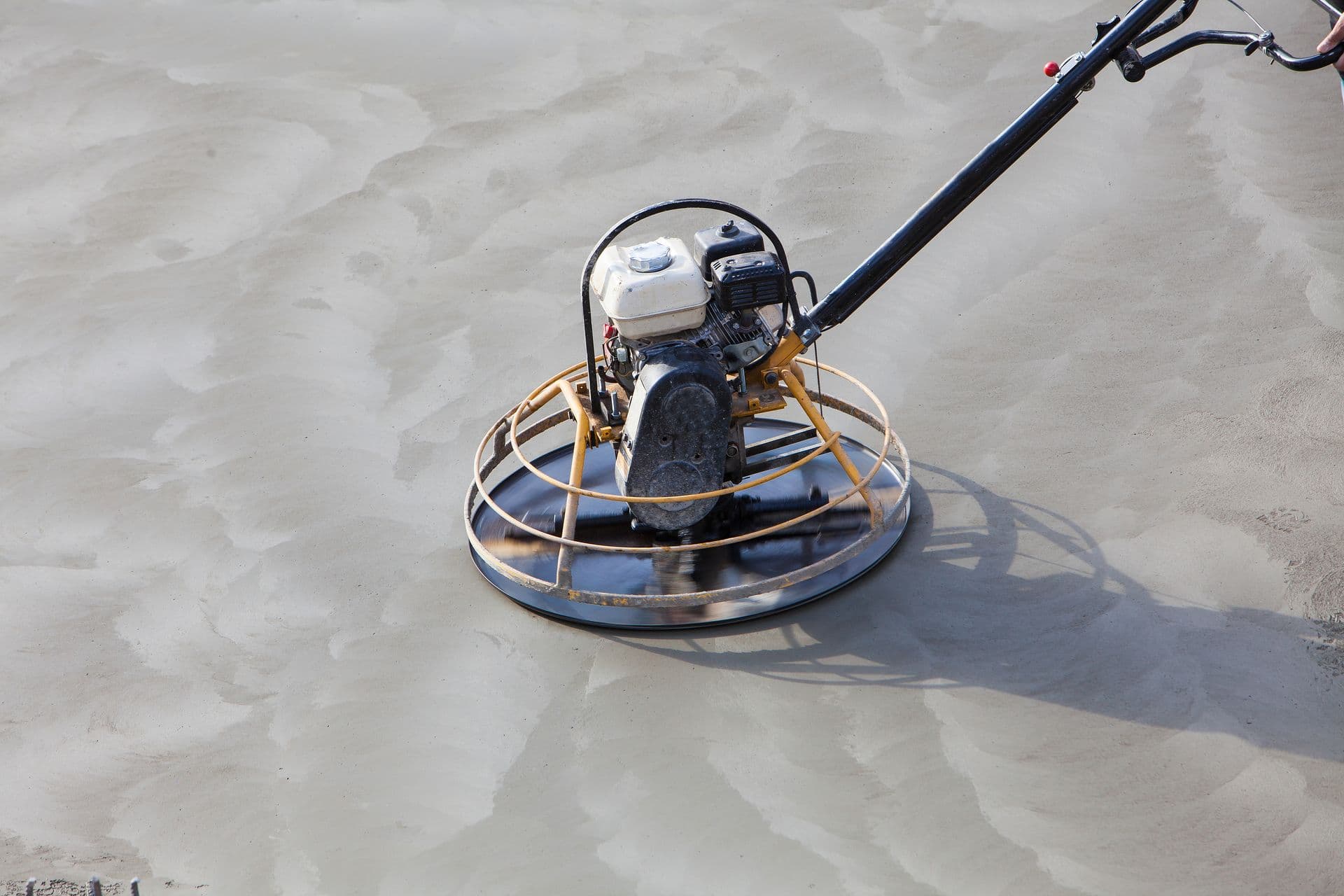 A concrete power trowel machine smoothing a wet cement surface. The metal blades create circular patterns, conveying a sense of motion and precision.