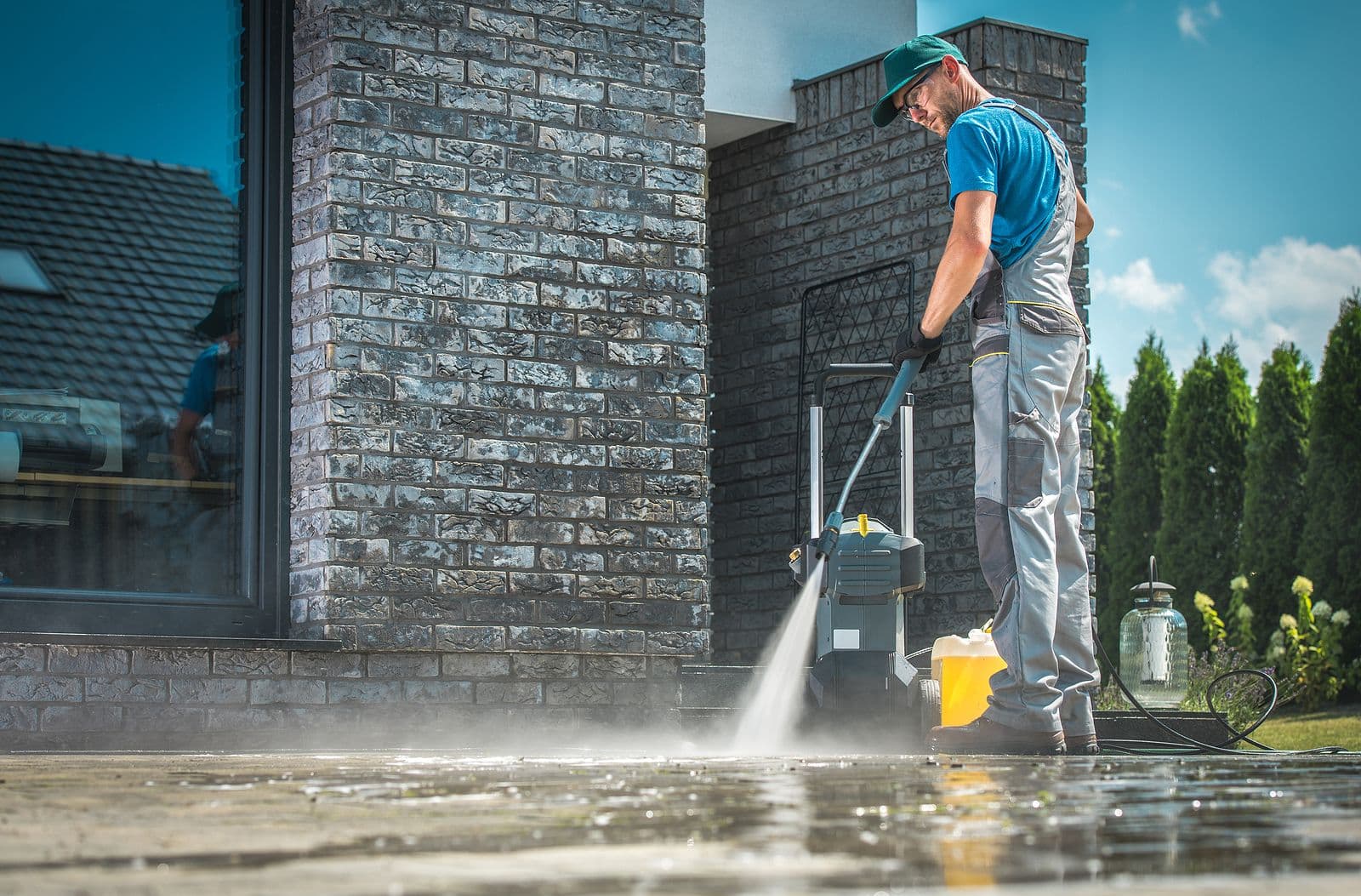 A person in work overalls and a cap uses a pressure washer to clean a modern stone patio. Bright blue sky and lush greenery enhance the fresh ambiance.