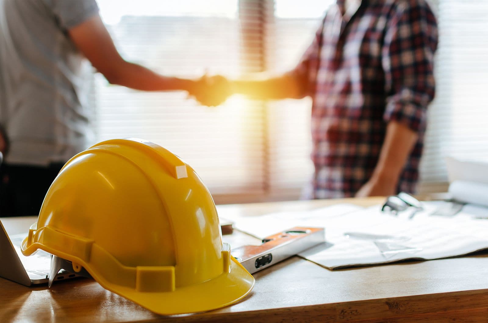 Yellow hard hat and plans on a desk in the foreground. Two people are shaking hands in the blurred background, conveying a successful agreement.