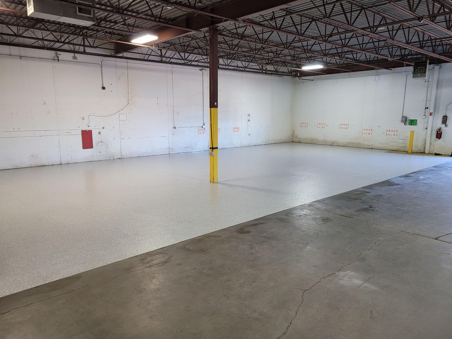 Empty warehouse interior with high ceilings, exposed beams, and a freshly coated gray speckled floor. A yellow support pillar stands in the center.