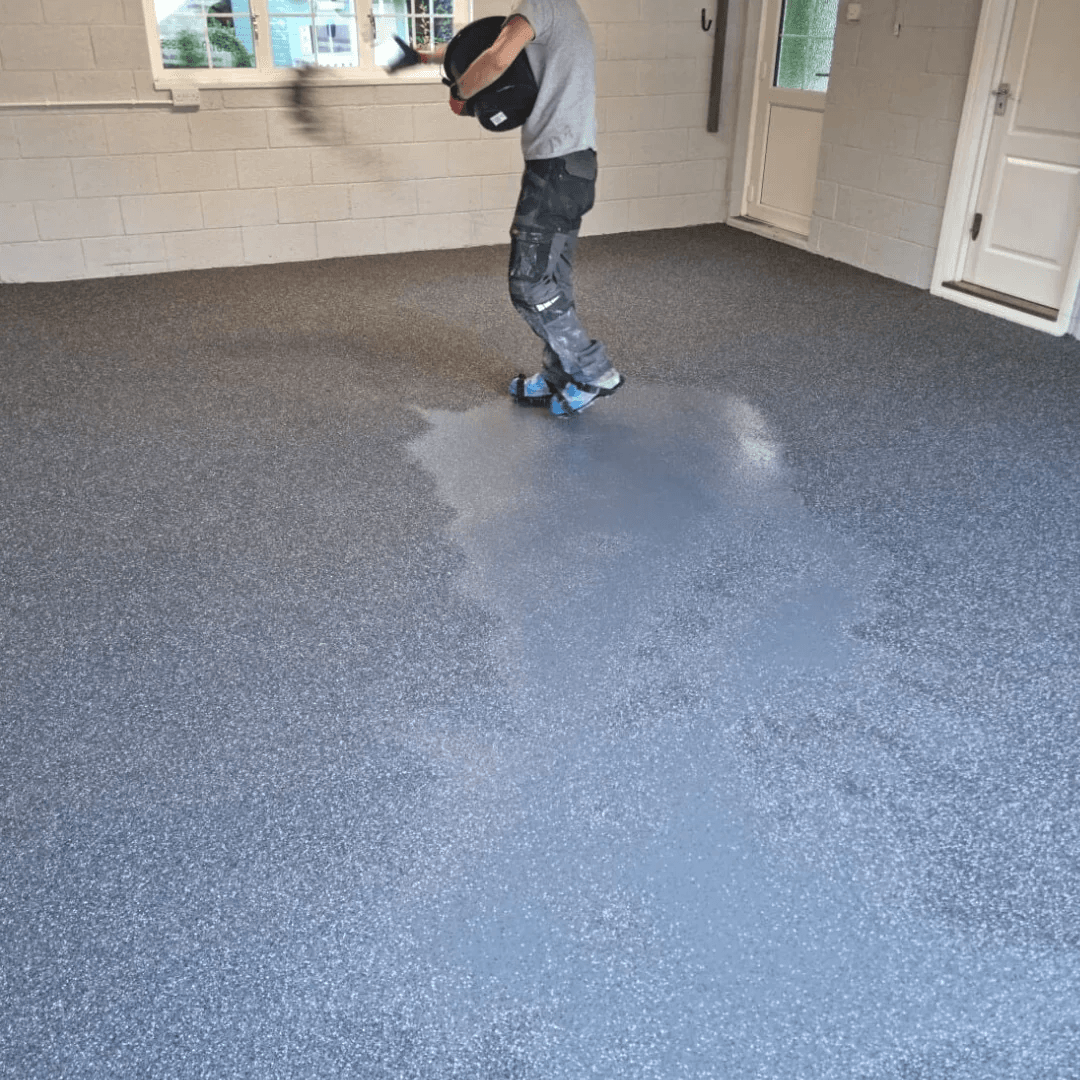 Worker in a gray shirt applies epoxy coating on a textured concrete floor in a garage. The setting is clean, organized, and well-lit.