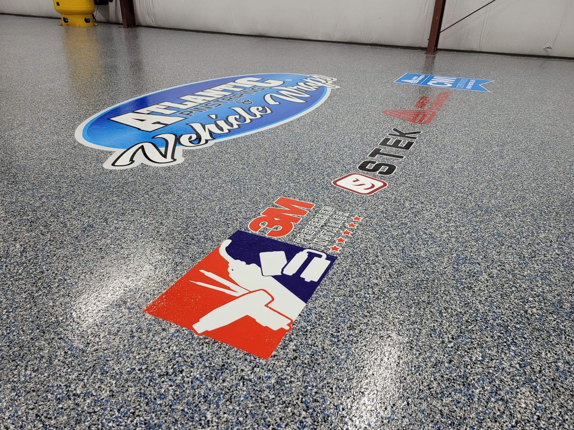 A speckled gray garage floor features vibrant logos, including a large blue and white oval with text. The scene conveys a professional atmosphere.