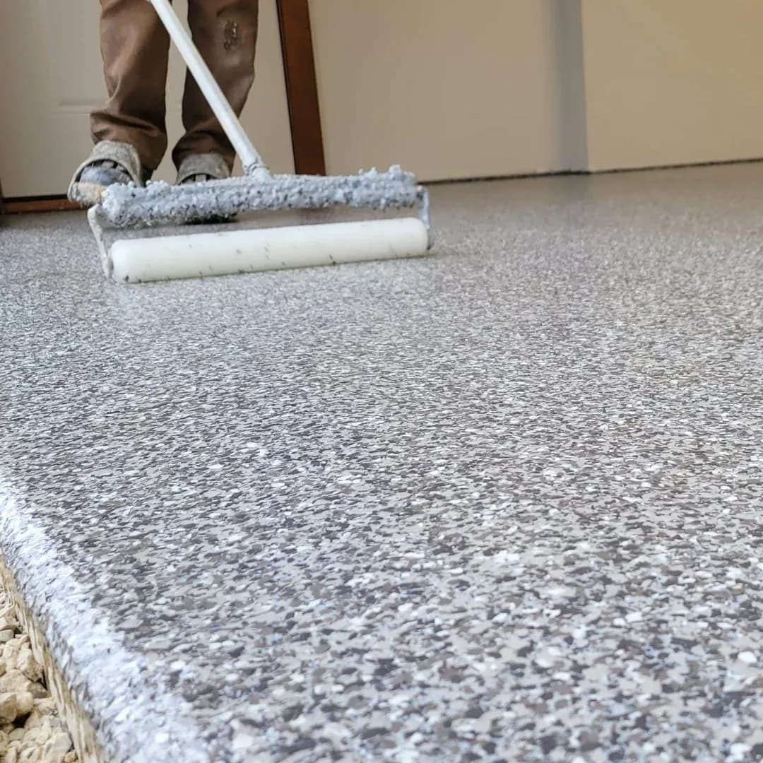 A person uses a roller to apply a gray speckled epoxy coating on a garage floor, creating a smooth, glossy finish. The scene conveys meticulous craftsmanship.