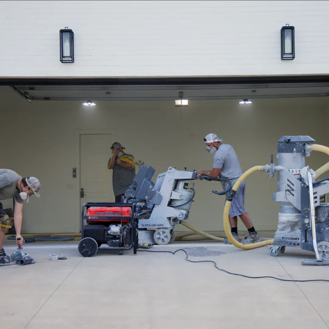 Workers smoothing concrete in a garage, using large industrial machines. They wear protective gear, focusing intently on tasks in a well-lit space.