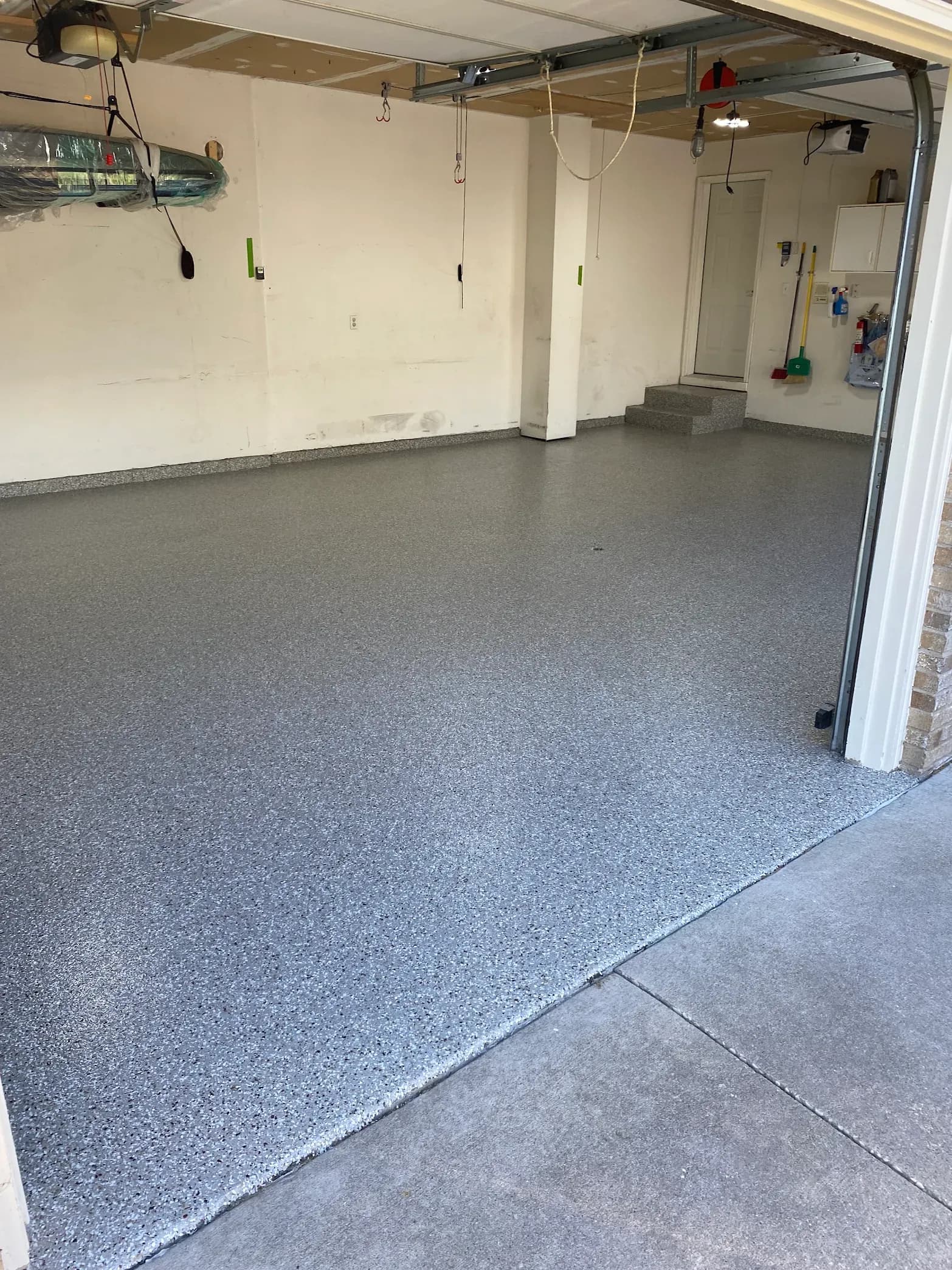 Empty garage interior with gray speckled epoxy flooring, white walls, central support column, overhead storage system with pulleys and hooks, and open garage door