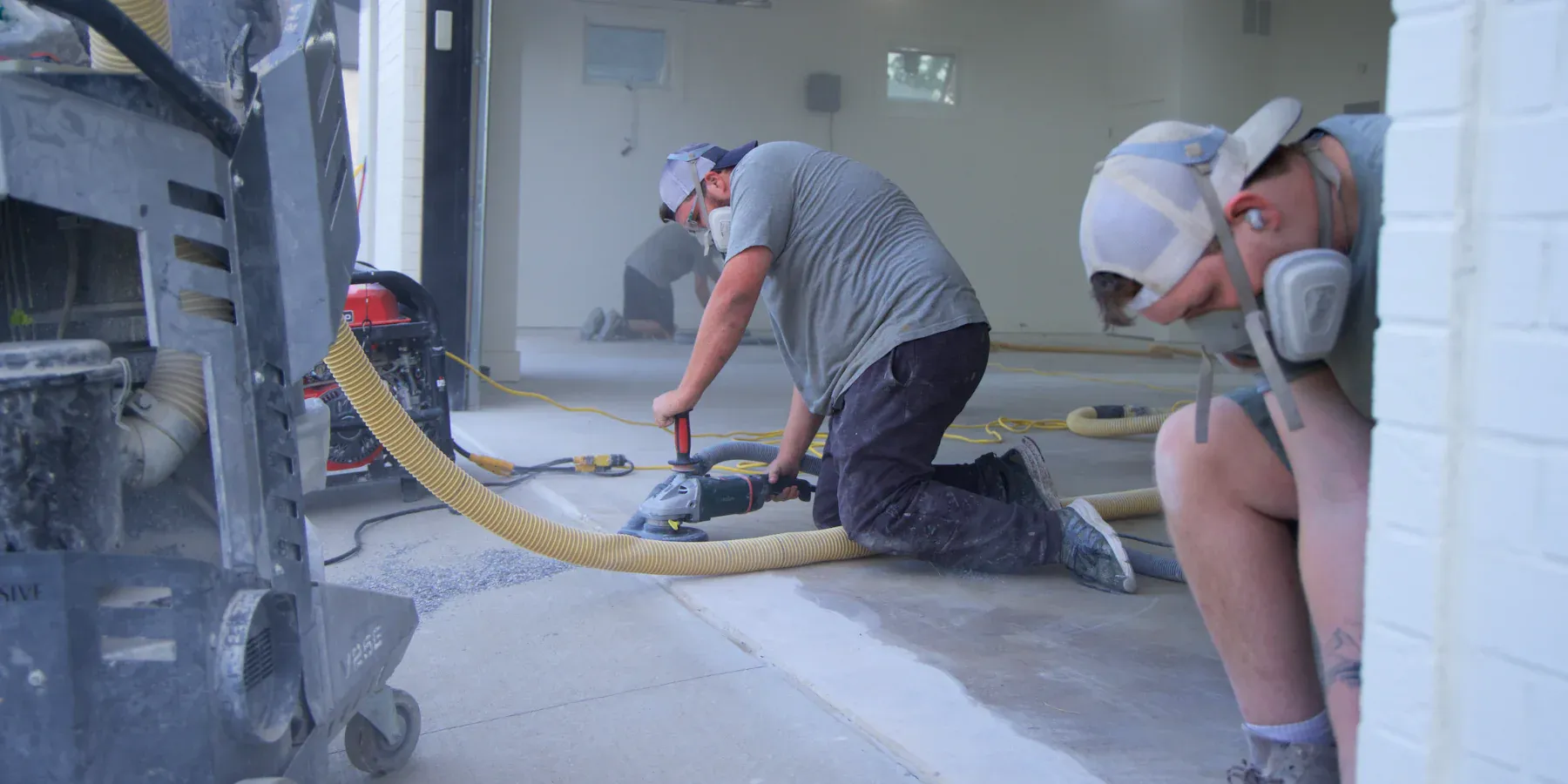 Two workers in protective gear are polishing a concrete floor in a garage. Dust fills the air as machinery runs. The scene conveys focus and diligence.